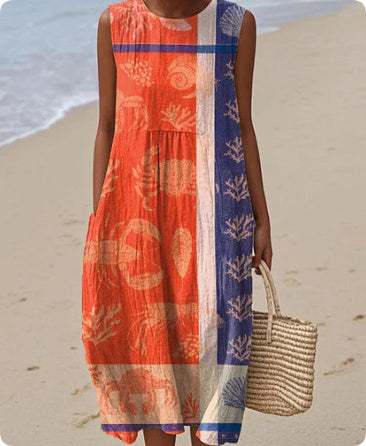 Woman in a red and blue dress with a pattern, holding a straw bag on a beach.